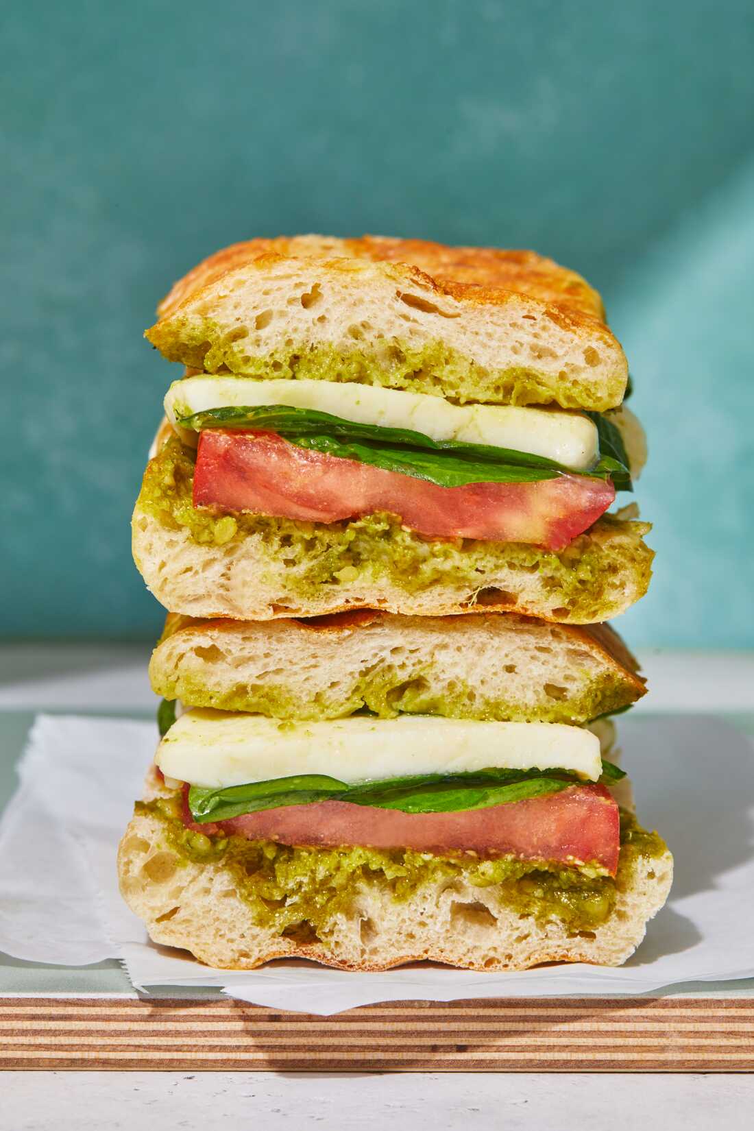 A close-up photo of a mozzarella, tomato and spinach pesto panini cut in half and wrapped on a wooden board in front of a watercolor background. 