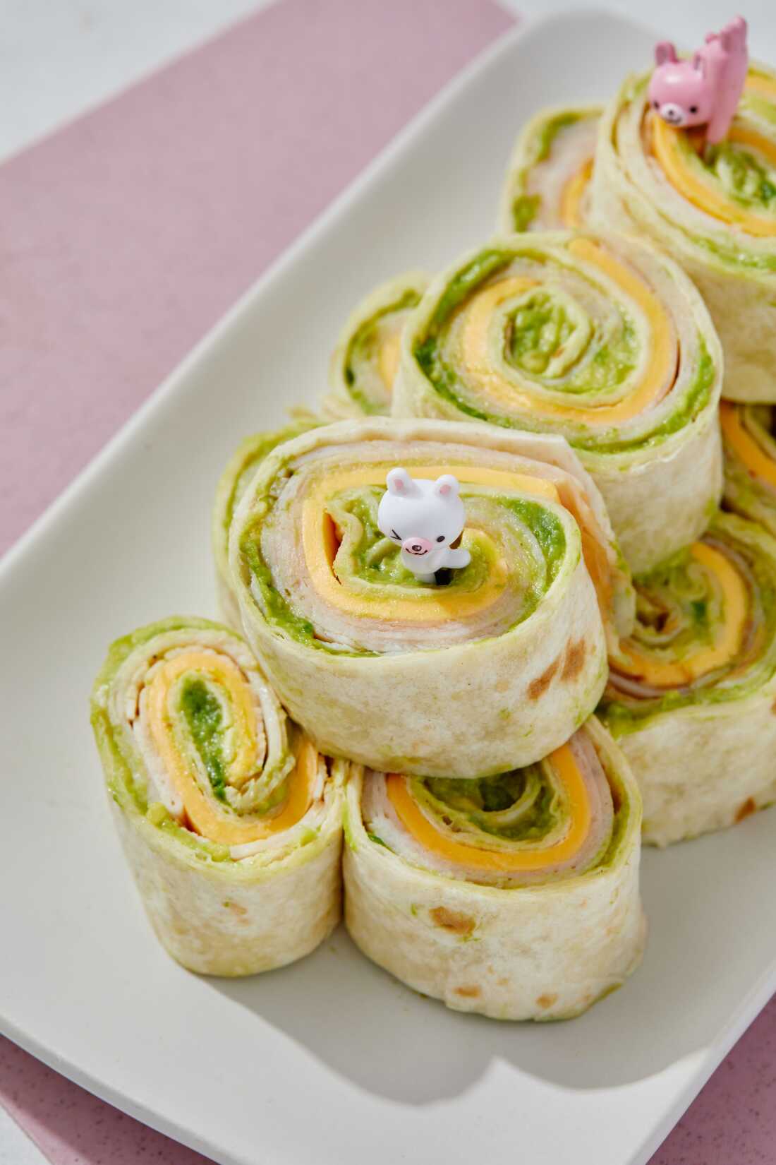 A photo of turkey, avocado and cheese pinwheels packed on a white plate. Chopped turkey, cheese and avocado are stuffed into a large flour tortilla and rolled up tightly.