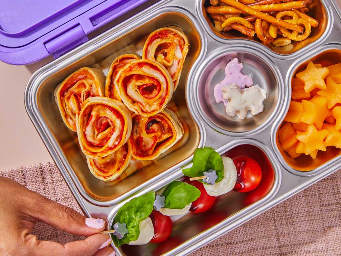 High resolution photo of purple and metal bento box lunchbox with pepperoni and mushroom pizza roll-ups, caprese salad bites, pretzels and fruit. 