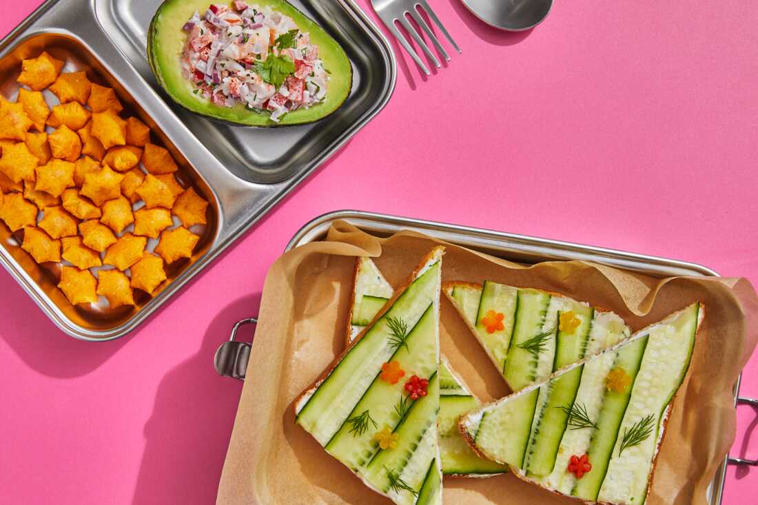 High angle view studio shot of an open-faced cucumber and avocado sandwich with shrimp in a metal box with pink sides. 